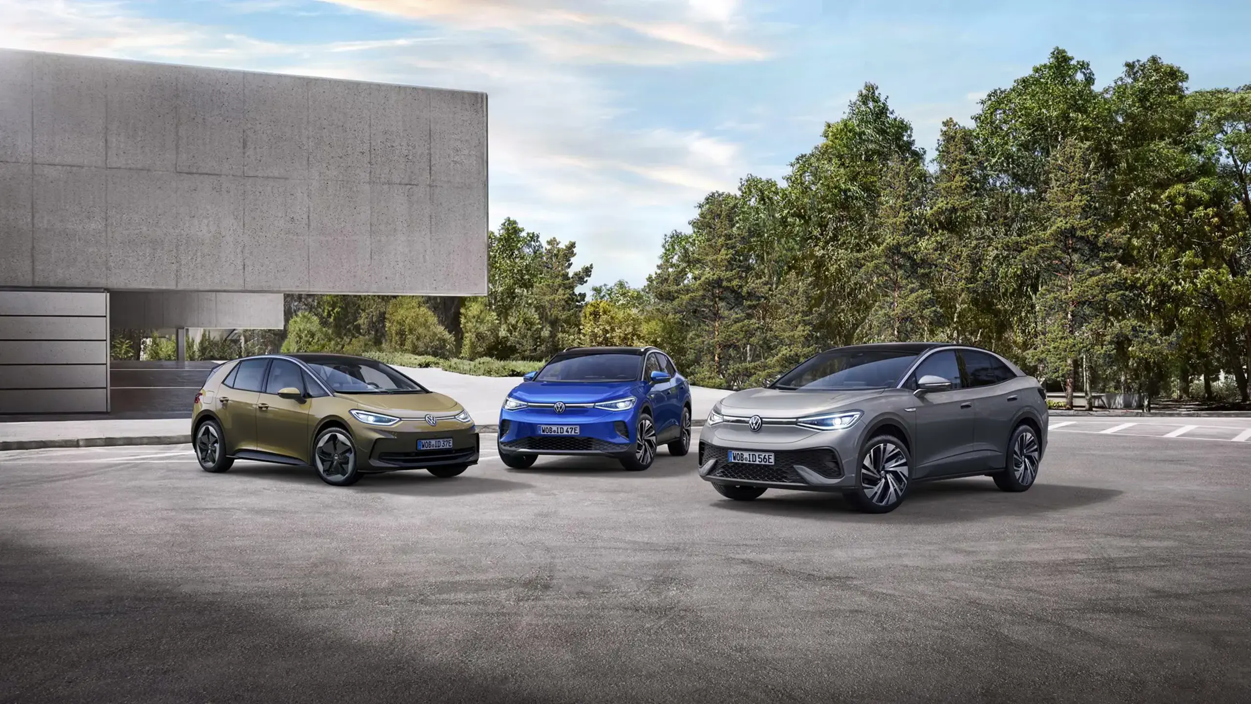 En grøn ID.3, Blå ID.4 og en grå ID.5 holder parkeret ved siden af hinanden ved en bygning med træer i baggrunden