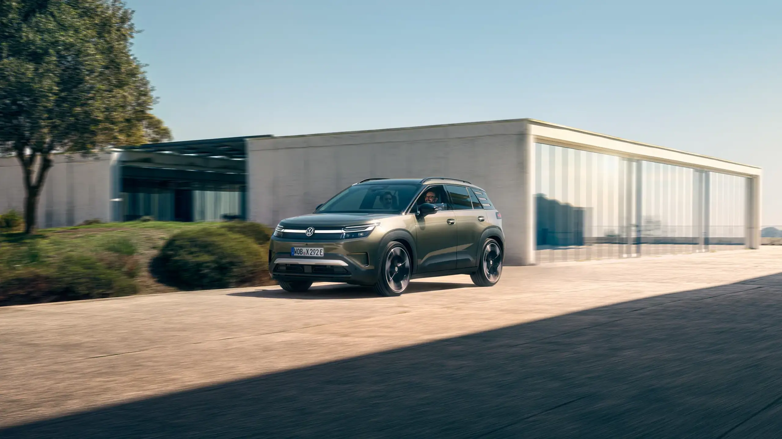 En grøn Volkswagen ID. Cross el-SUV ses forfra fra førersiden, mens den kører foran en moderne bygning på en solrig dag.